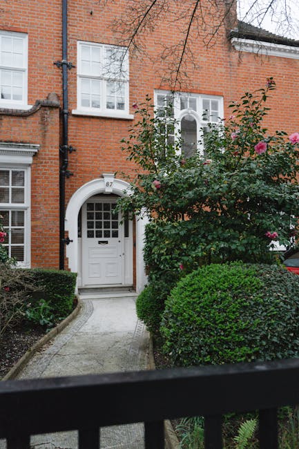 A view of a residential terraced garden in an urban setting, featuring a narrow paved pathway leading to a white front door with glass panes, set within a red-brick house. The garden is well-maintaine